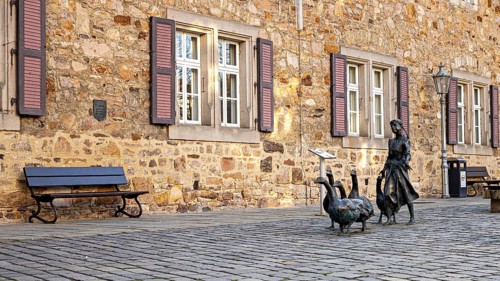 Scene in front of a stone building with shutters, a bench and a statue of a girl with geese, The