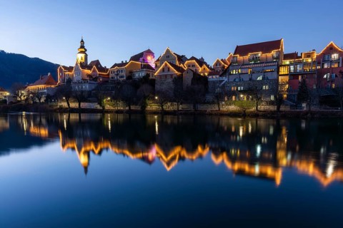Christmas illuminated house facade of Frohnleiten reflected in the river Mur, blue hour,