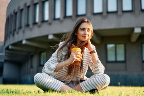 Young stylish woman drinking coffe and relax sitting on a grass in city park. Break time concept