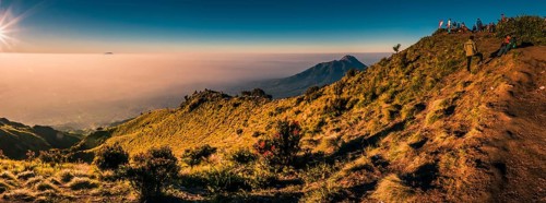 Panoramic photo of mountains surrounding Mount Merbabu and hikers at top during sunrise near Yogya