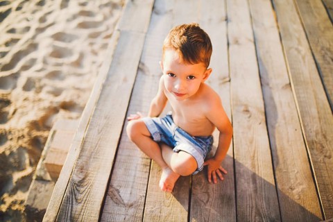 Little Caucasian boy child sitting on wooden pier sandy beach, summer time, sea holiday near water.