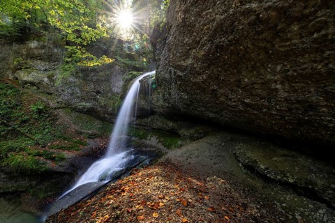 A waterfall flows over mossy rocks, illuminated by sunbeams breaking through the leaves,