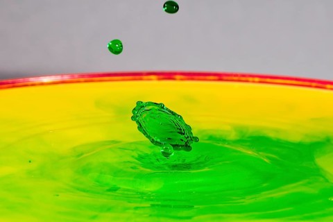 A green drop hits a yellow-green liquid and creates colourful patterns, water droplets, collision,