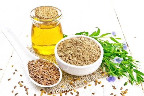 Flaxseed flour in a bowl, linenseeds in a spoon, oil in glass jar on burlap napkin, leaves and blue