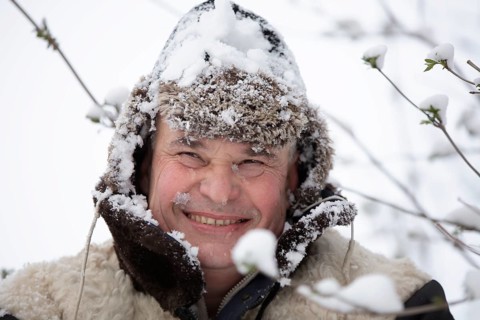 An elderly man in a snowy winter hat smiles and looks into the camera