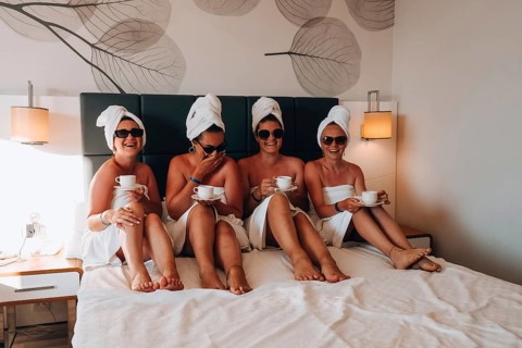 Four girls friends in white towels sitting on the bed having fun in hotel. Vacation Bloggers