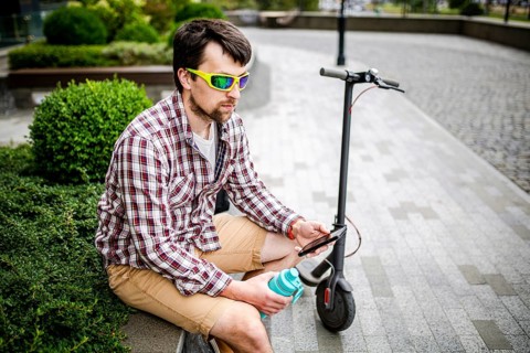 Man with e-scooter relaxing and using smartphone on bench in city. Modern male with electro scooter