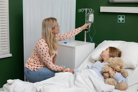 In a hospital, a caring mother supports her child with an IV, who holds a beloved teddy bear for
