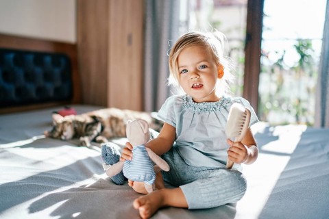 Little smiling girl sitting on the bed with a teddy bear and a comb in her hands.
