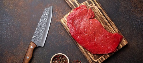 Raw uncooked top round beef steak on wooden cutting board with big kitchen knife and pepper on dark