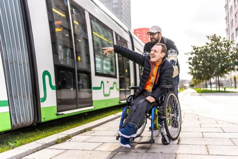 Disabled man in a wheelchair points at something while his friend stands behind him near a tram