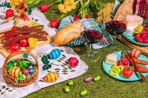 A picnic with two wine glasses on grass, surrounded by bread, sausage, cheese, vegetables and