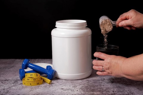 Woman pouring protein powder into a shaker with a spoon, next to a bottle of protein, dumbbells and