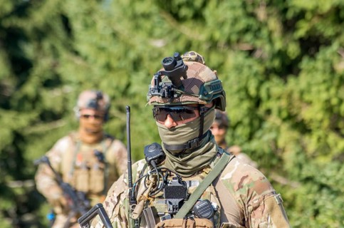 A soldier in camouflage uniform with tactical gear and sunglasses patrols a forest area