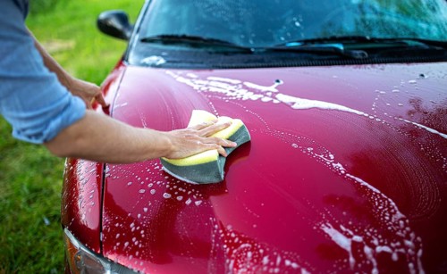 Hand using a sponge to wash the hood of a red car with soap and suds