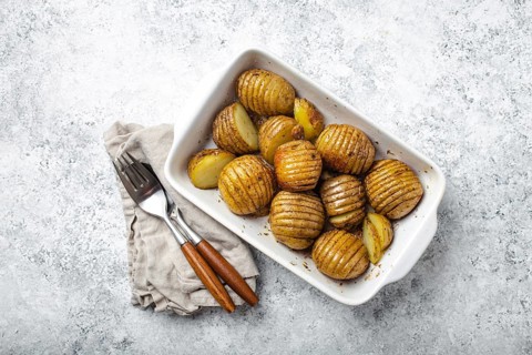 Hasselback potatoes with herbs in white ceramic casserole dish on white concrete table background