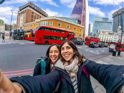 Two happy female tourists taking a selfie with red double decker buses and modern skyscrapers in