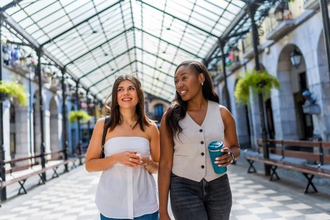 Two young businesswomen are walking and talking in a covered gallery, enjoying a break and holding