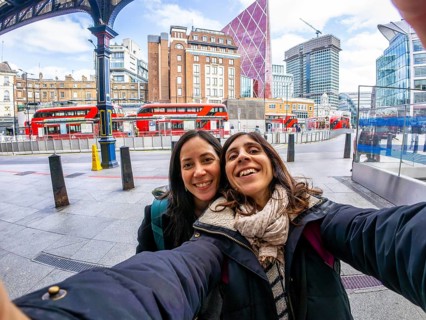 Two happy tourists taking a selfie in the city of london, with red buses and modern buildings in