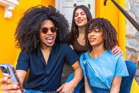 Three happy young friends are taking a selfie with a smartphone while sitting on the stairs of a