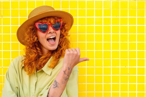 Excited young woman with red hair, heart shaped sunglasses, and straw hat pointing her thumb to the