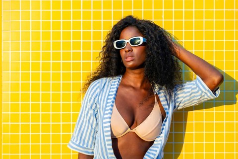 Stylish model with sunglasses and striped shirt posing against a vibrant yellow tiled wall,