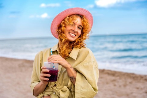 Young woman with curly red hair and pink hat enjoying a refreshing cold beverage on a sunny beach