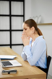 A business woman sits at her desk with a laptop, hands clasped in front of her mouth, visibly deep