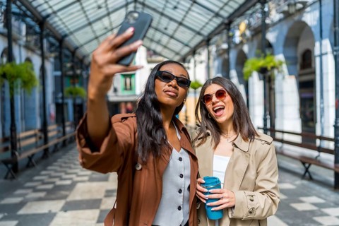 Two women are taking a selfie with a smartphone while walking down the street, holding reusable