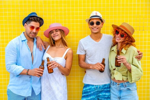 Four cheerful friends wearing summer clothes and hats are enjoying a refreshing beer against a