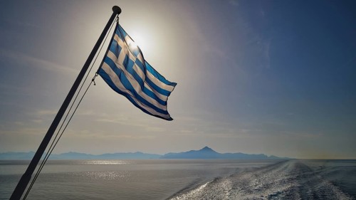 Greek flag in the foreground, over a widening sea under a blue sky, crossing Rhodes to Symi, ferry,