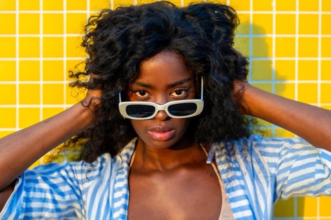 Fashionable model with curly hair touching her head while wearing white sunglasses and posing in