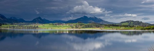 Panorama at sunrise over Lake Hopfensee to the sunlit western shore with farms, behind it receding