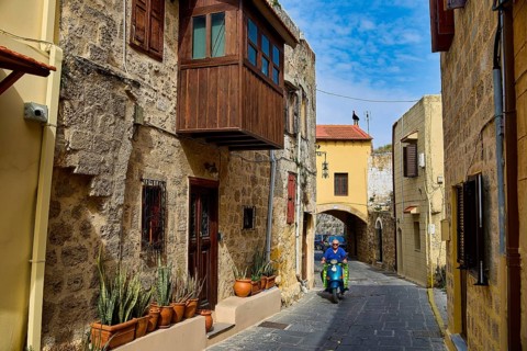 Narrow historic alley with a scooter driver and flower pots on a stone building, Rhodes Old Town,