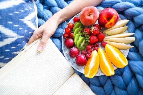 Plate with fresh fruit salad on a blue plaid and book. Woman reading the book and eating fresh