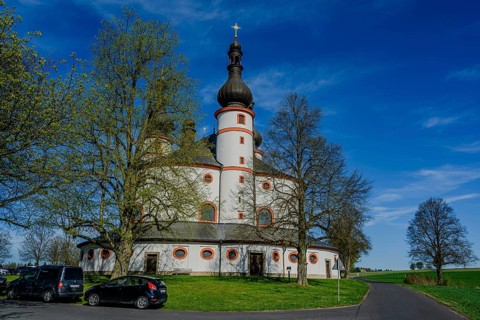 A baroque church with round architecture under a clear blue sky, surrounded by trees and a road,