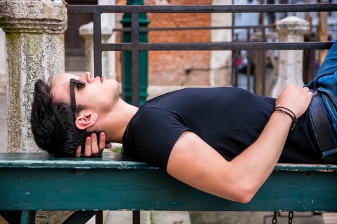 Attractive young man laying down on wood bench in European city, wearing sunglasses