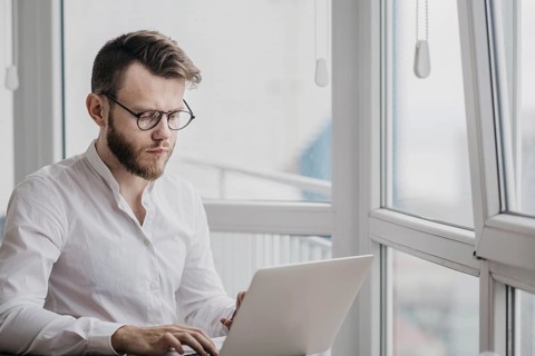 Man in a white shirt and glasses focused on his laptop in a bright room