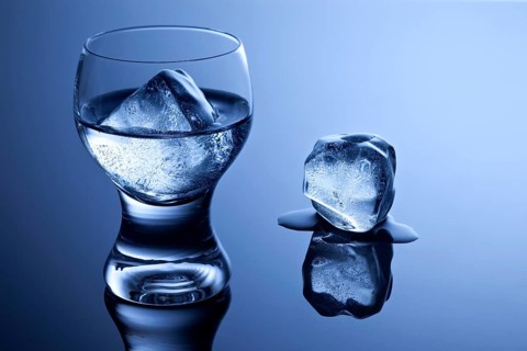 An elegant shot glass with vodka and ice and a melting ice cube on a reflective surface