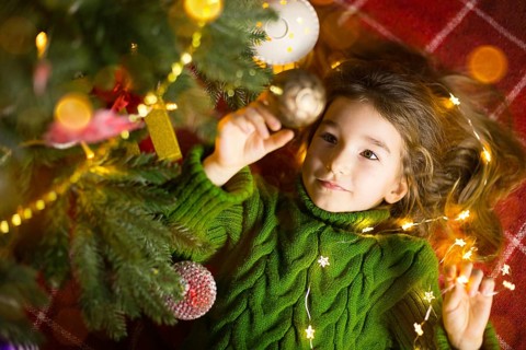 A girl with long hair and garlands lies on a red plaid under a Christmas tree with toys in a warm