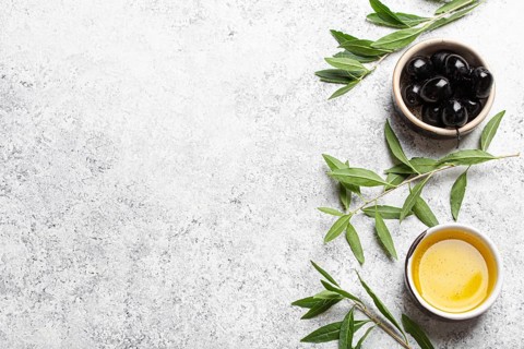 Black olives and extra virgin olive oil in little bowl with olive tree branches top view on white