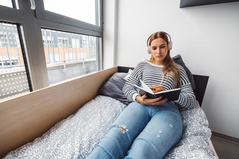 Blonde young caucasian woman lying on her dorm room bed next to a window putting on headphones,