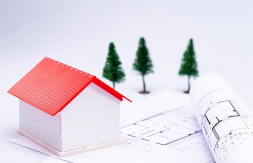 The photo shows a model house with architect's plans, in the background you can see some fir trees