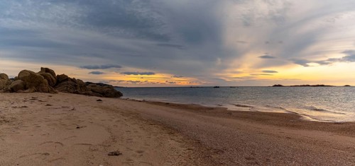 A panorama view of a golden sand beach at sunset with calm ocean water and red granite rocks
