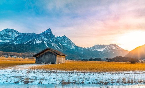 Winter alpine landscape with an aged wooden barn, the Alps mountains, a frozen river at sundown.