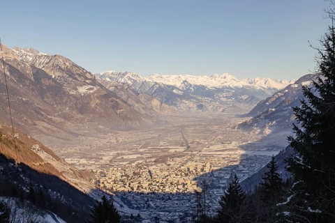 A valley town surrounded by snowy mountains at sunset, casting long shadows over the landscape,