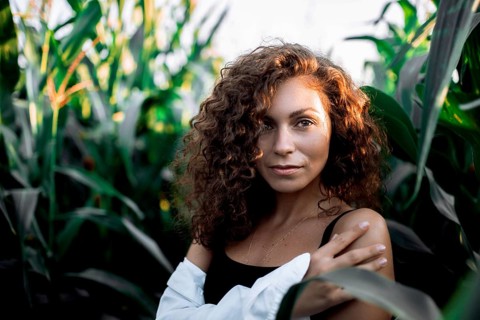 A confident woman with curly hair stands among corn stalks, crossing her arms