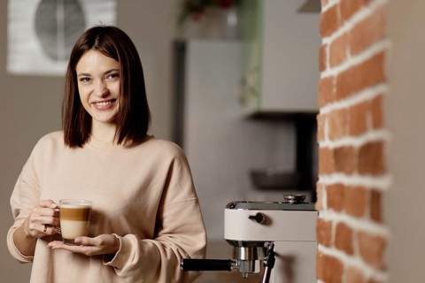 Woman with a cup of coffee beside an espresso machine and brick wall