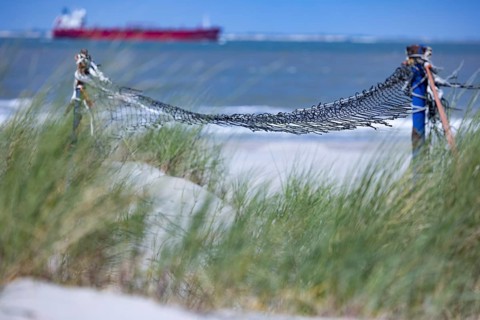 Old, weathered volleyball net in front of grasses on the beach, a ship can be seen in the distance,