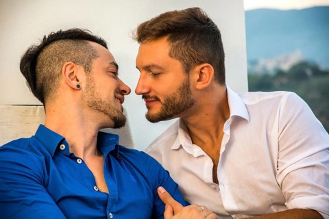 Two gays sitting on chairs at balcony holding hands and looking at each other